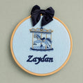 Ice blue silk name frame in a light wooden hoop with a navy satin bow, an embroidered carousel with gold canopy and a name in navy script