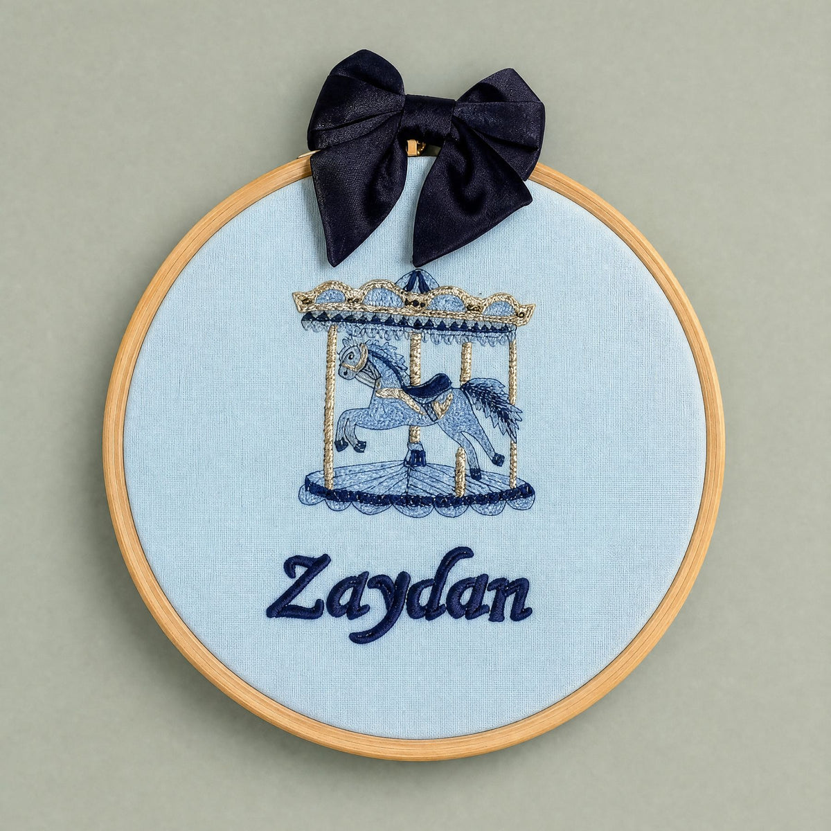 Ice blue silk name frame in a light wooden hoop with a navy satin bow, an embroidered carousel with gold canopy and a name in navy script