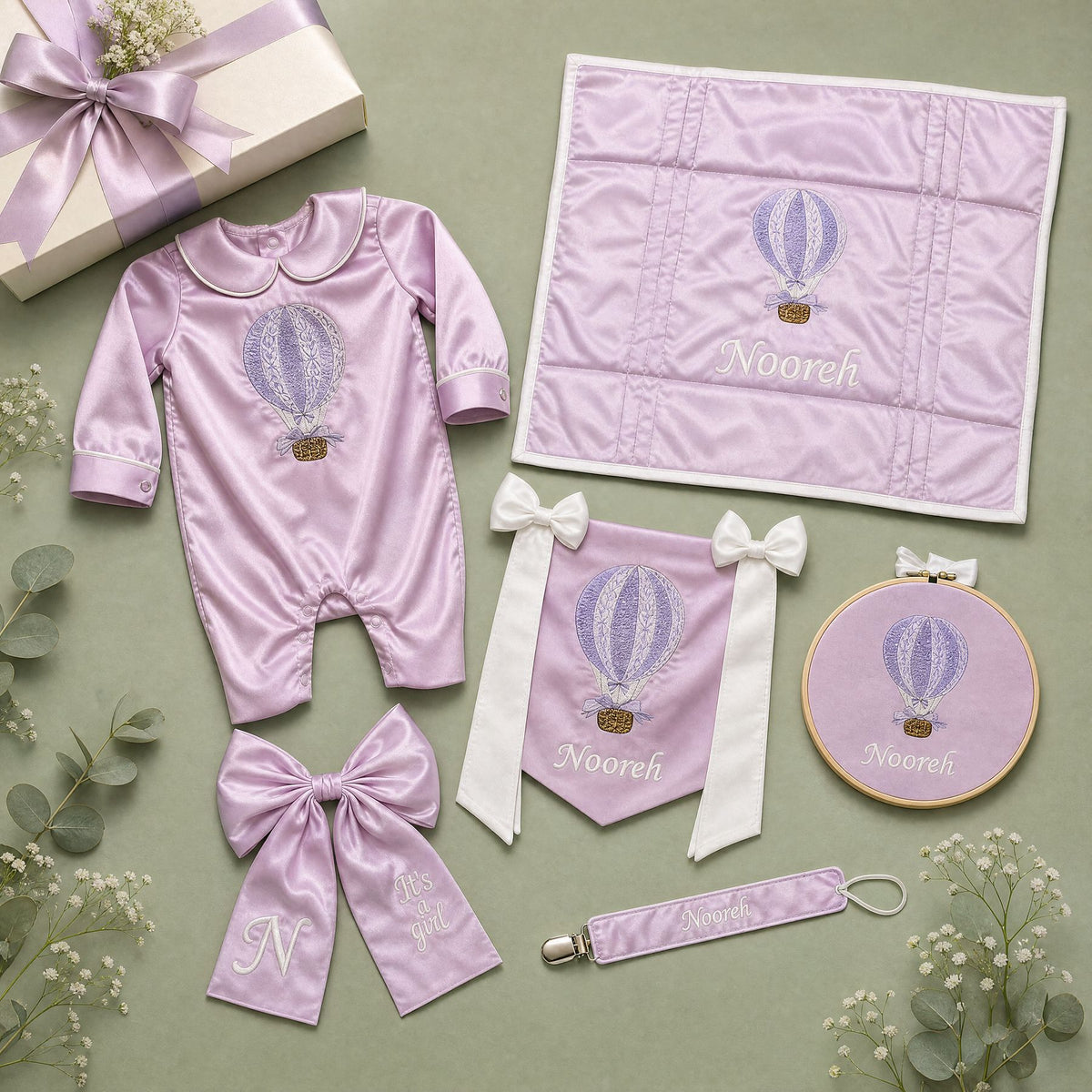 The Grand Arrival lilac gift bundle laid out flat — all six personalised lilac silk pieces including romper, wrapping sheet, name frame, name flag, announcement bow and pacifier holder
