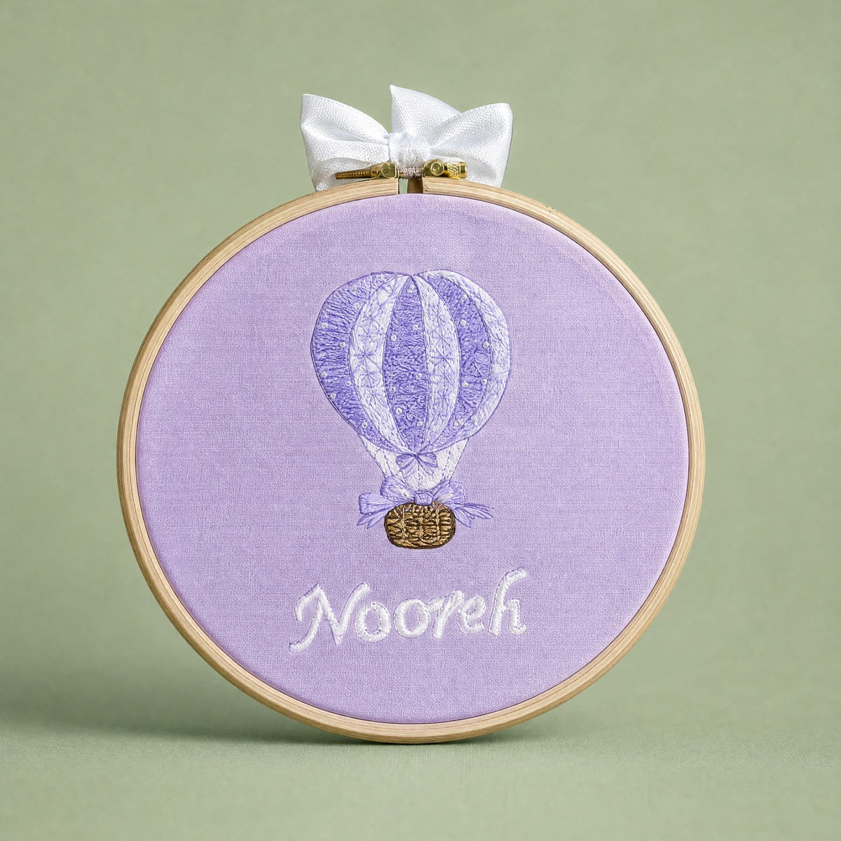 Lilac silk name frame in a light wooden hoop with an ivory satin bow, an embroidered hot air balloon and a name in ivory script