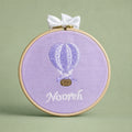 Lilac silk name frame in a light wooden hoop with an ivory satin bow, an embroidered hot air balloon and a name in ivory script