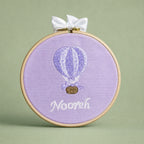 Lilac silk name frame in a light wooden hoop with an ivory satin bow, an embroidered hot air balloon and a name in ivory script