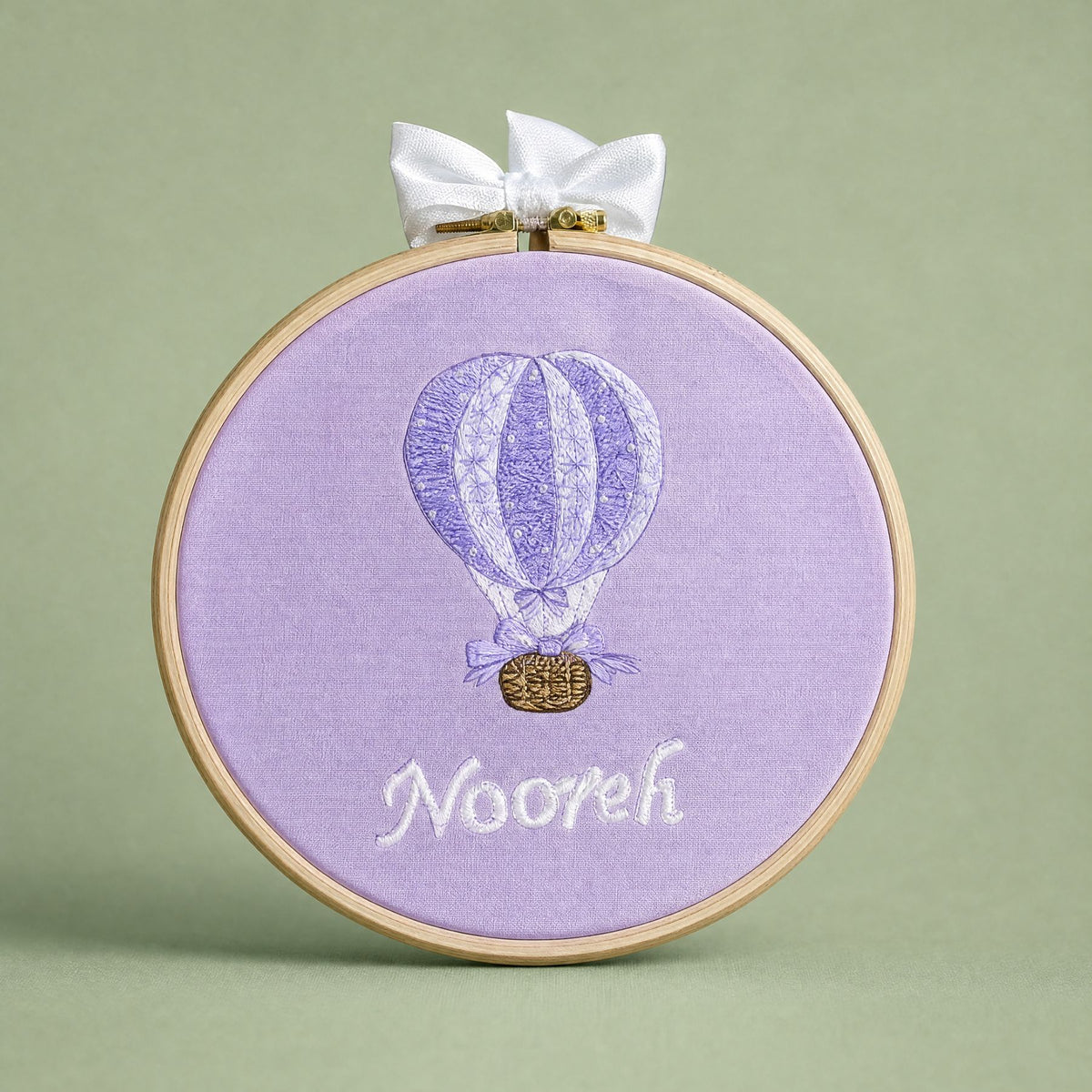 Lilac silk name frame in a light wooden hoop with an ivory satin bow, an embroidered hot air balloon and a name in ivory script