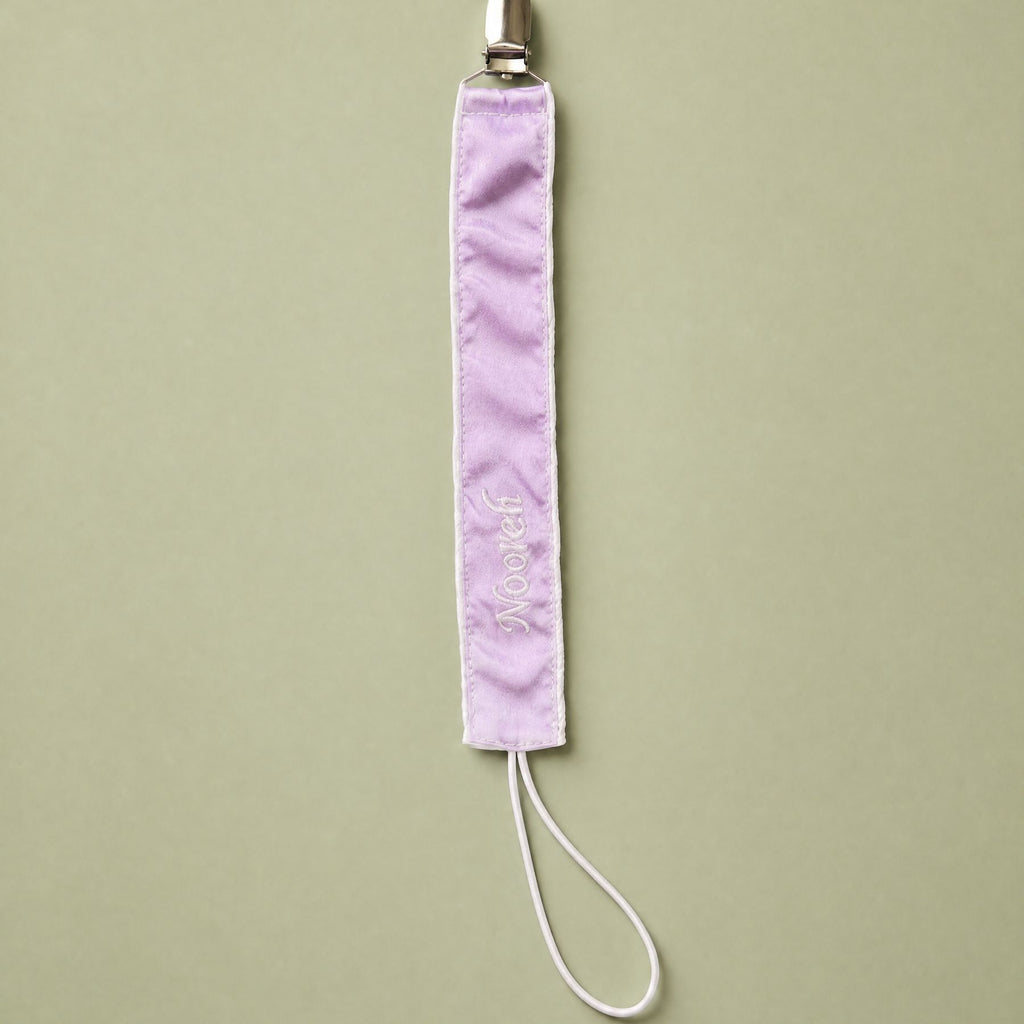 Lilac silk pacifier holder with ivory detailing and a baby's name embroidered in ivory script
