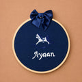 Navy silk name frame in a light wooden hoop with a navy satin bow, an embroidered rocking horse and a name in ivory script