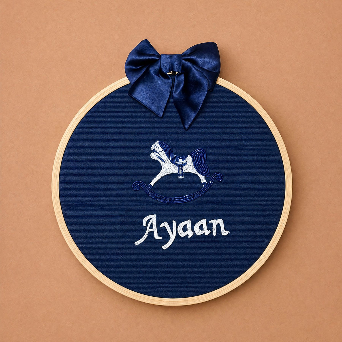 Navy silk name frame in a light wooden hoop with a navy satin bow, an embroidered rocking horse and a name in ivory script