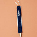 Navy silk pacifier holder with ivory piping, a gold clip and a name embroidered in ivory script