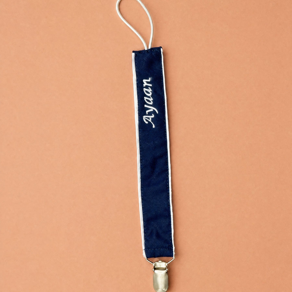 Navy silk pacifier holder with ivory piping, a gold clip and a name embroidered in ivory script