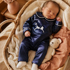 Newborn sleeping in a woven moses basket wearing the navy silk Zyra Baby romper, beside a knitted teddy