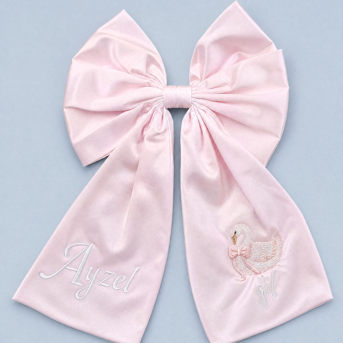 Large soft pink silk announcement bow with a baby's name in ivory on one tail and an embroidered swan with It's a girl on the other