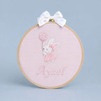 Pink embroidery hoop name frame with a bunny holding a flower-printed balloon, a white satin bow and a baby's name in pink script