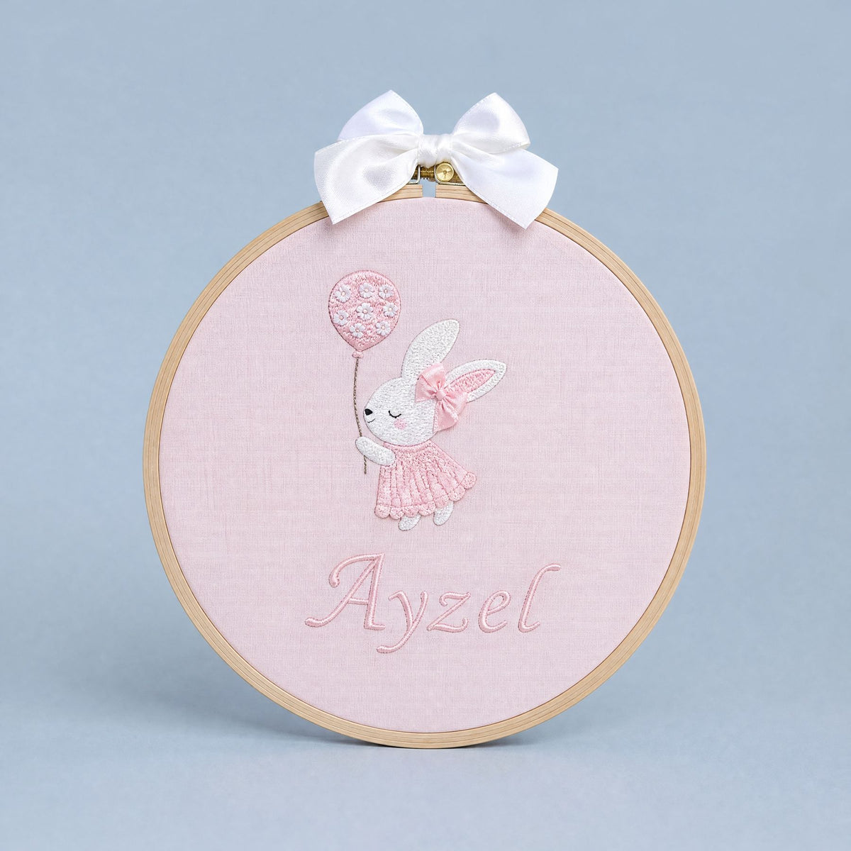 Pink embroidery hoop name frame with a bunny holding a flower-printed balloon, a white satin bow and a baby's name in pink script