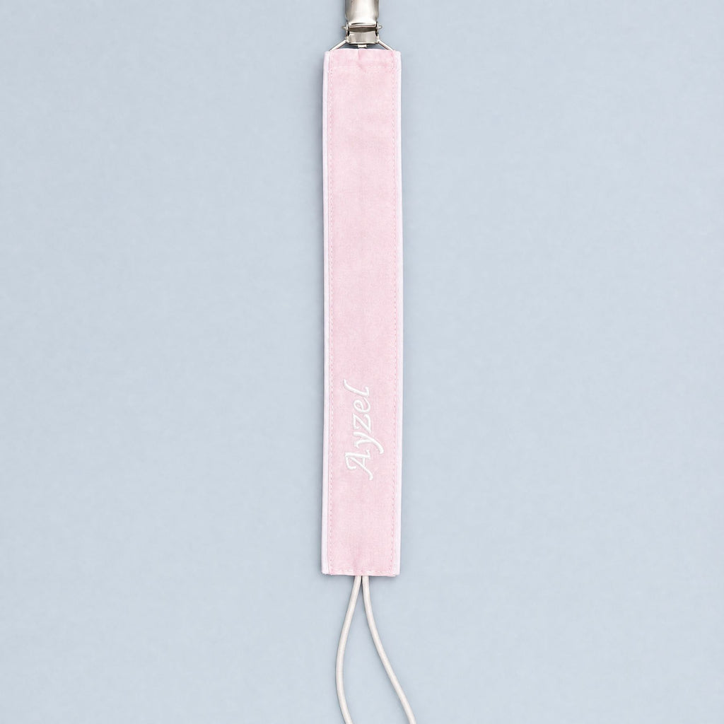 Soft pink silk pacifier holder with white edging, a metal clip, ivory cord loop and a baby's name embroidered in ivory script