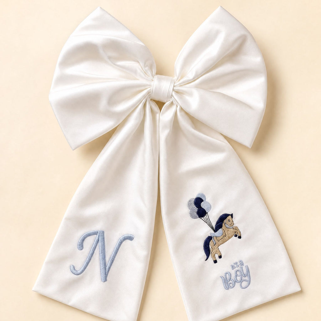 Oversized ivory silk announcement bow with a pale blue embroidered initial on one tail and a pony holding balloons with It's a Boy lettering on the other