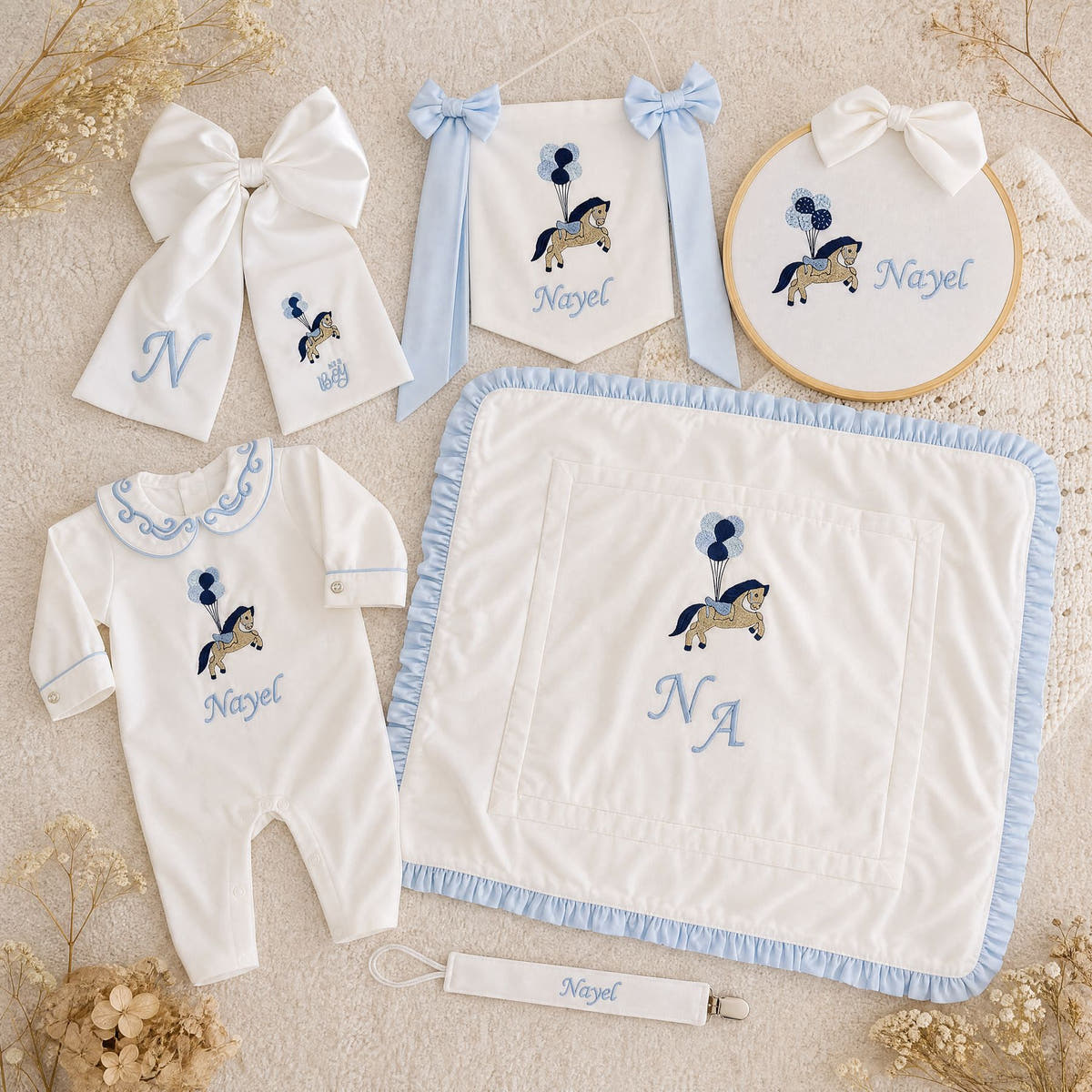 The Grand Arrival white and blue gift bundle laid out flat — all six personalised ivory silk pieces including romper, wrapping sheet, name frame, name flag, announcement bow and pacifier holder