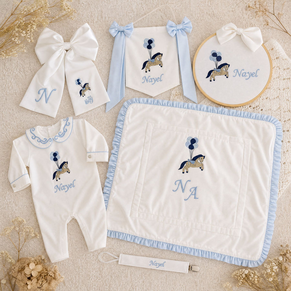 The Grand Arrival white and blue gift bundle laid out flat — all six personalised ivory silk pieces including romper, wrapping sheet, name frame, name flag, announcement bow and pacifier holder