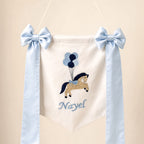 Ivory silk name flag with an embroidered pony and balloons, silk bows and a baby's name in pale blue script
