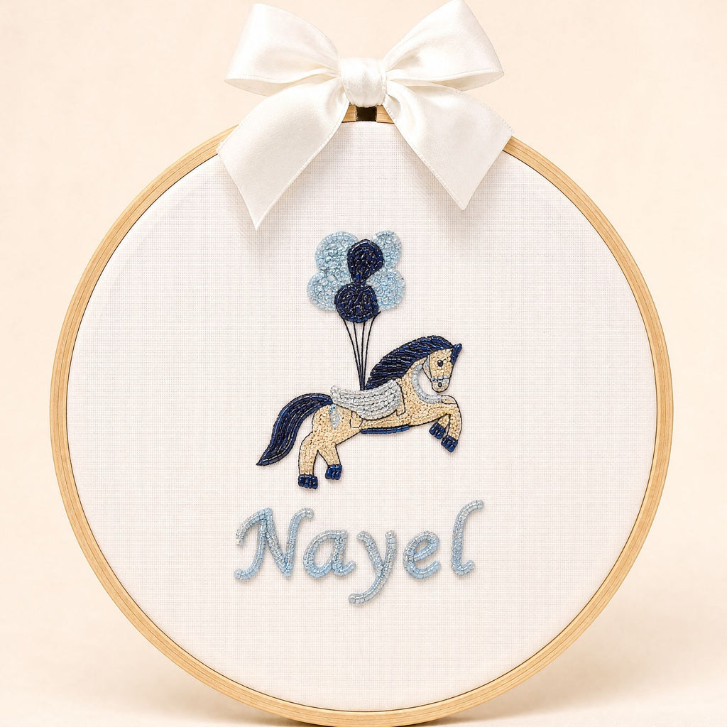 Ivory silk name frame in a light wooden hoop with an embroidered pony and balloons and a name in pale blue script