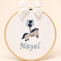 Ivory silk name frame in a light wooden hoop with an embroidered pony and balloons and a name in pale blue script