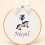 Ivory silk name frame in a light wooden hoop with an embroidered pony and balloons and a name in pale blue script