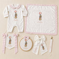 The Grand Arrival white and pink gift bundle laid out flat — all six personalised ivory silk pieces including romper, wrapping sheet, name frame, name flag, announcement bow and pacifier holder