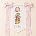 Ivory silk name flag with an embroidered bunny holding a pink balloon, soft pink silk bows and a baby's name in pink script