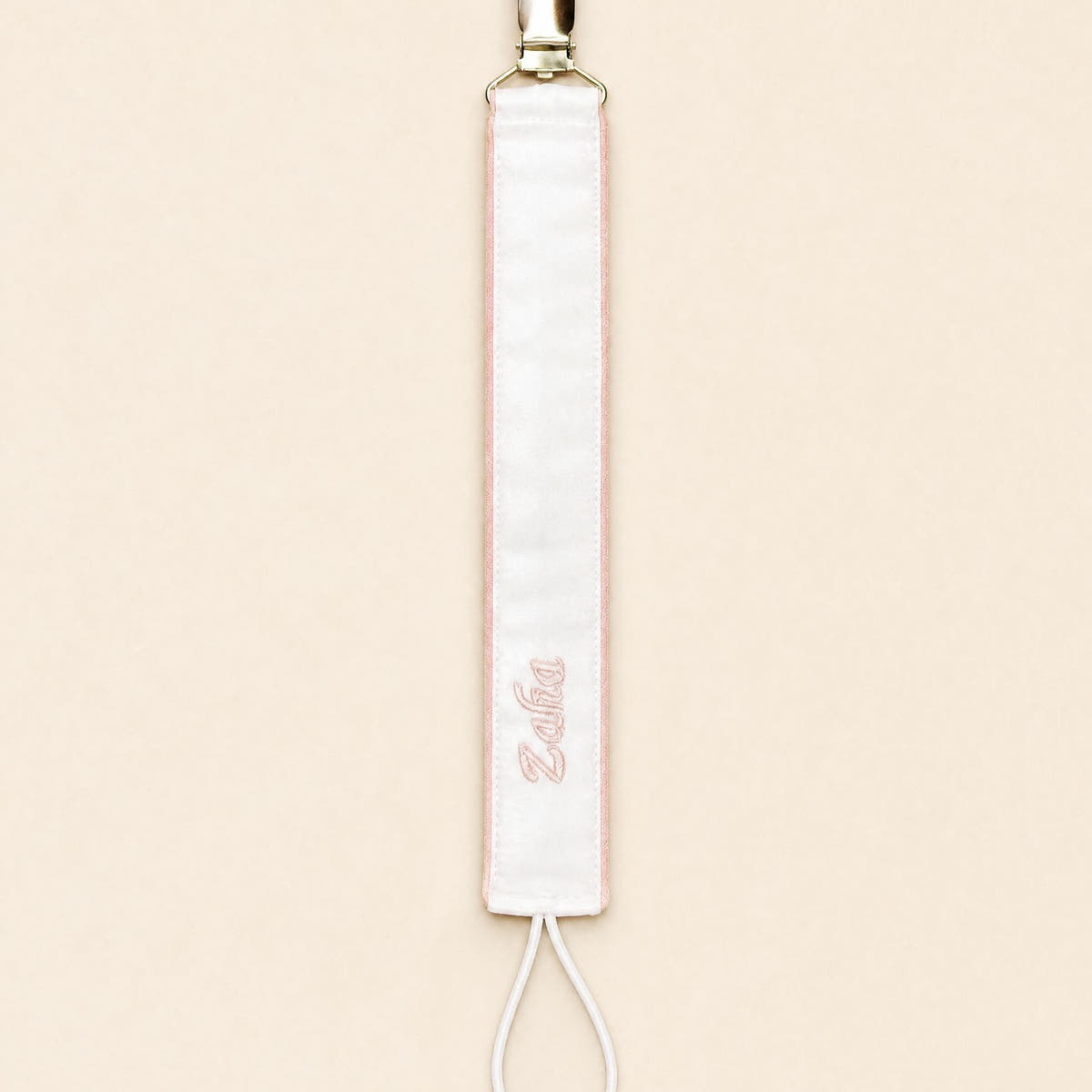 Ivory silk pacifier holder edged in soft pink with a gold clip, cord loop and a baby's name embroidered in pink script