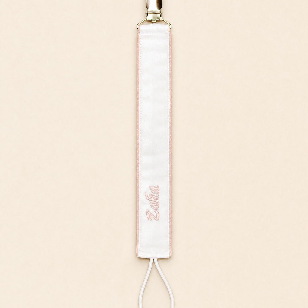 Ivory silk pacifier holder edged in soft pink with a gold clip, cord loop and a baby's name embroidered in pink script
