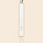 Ivory silk pacifier holder edged in soft pink with a gold clip, cord loop and a baby's name embroidered in pink script
