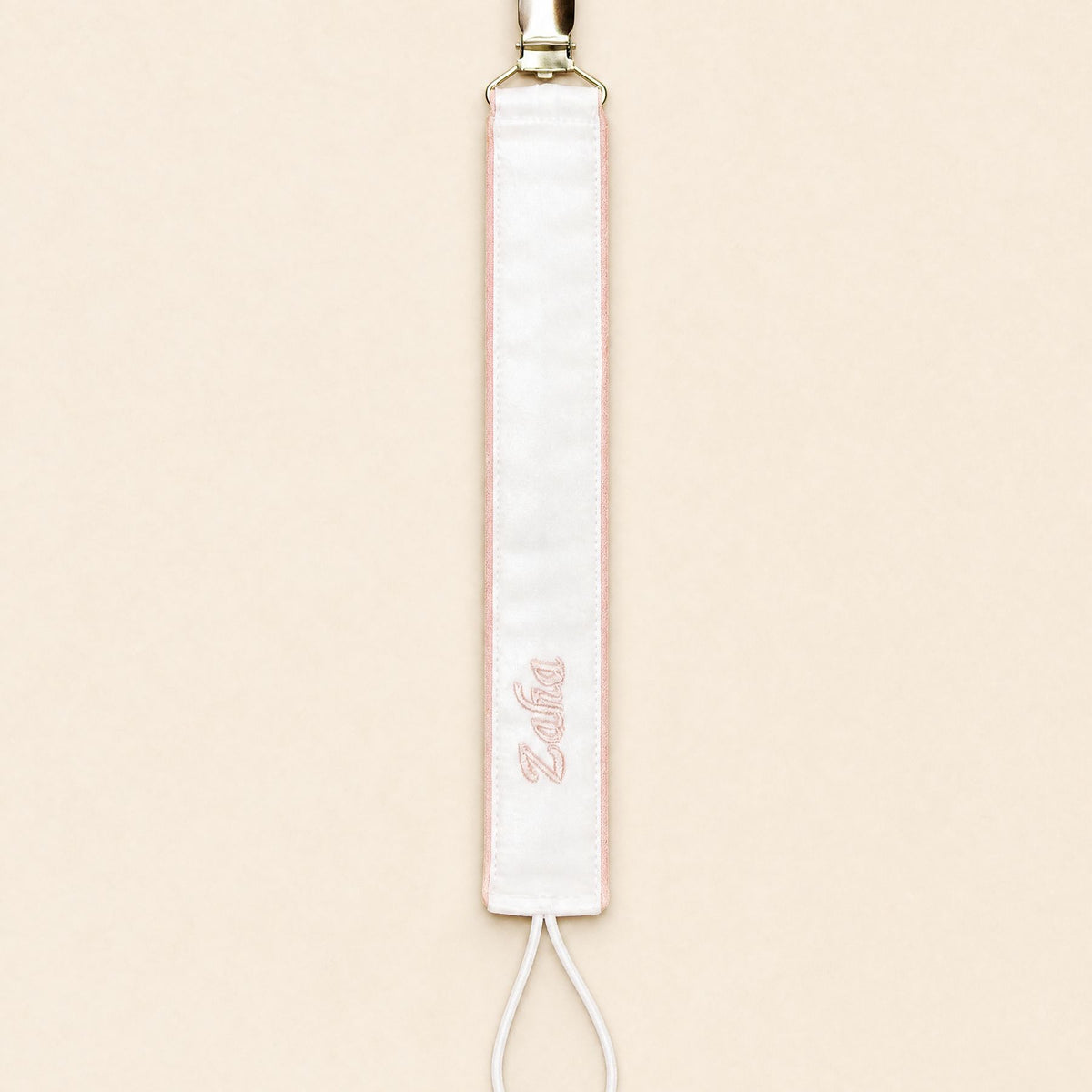 Ivory silk pacifier holder edged in soft pink with a gold clip, cord loop and a baby's name embroidered in pink script